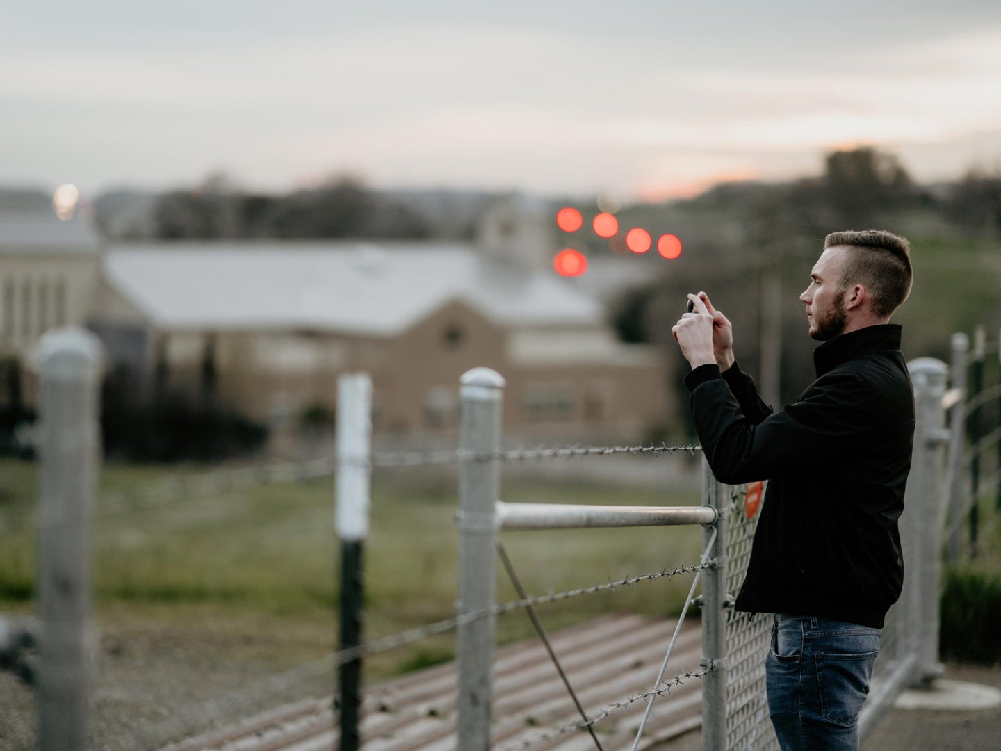 A man standing by a fence, taking a photo on an iPhone.