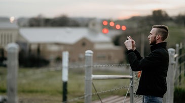 A man standing by a fence, taking a photo on an iPhone.