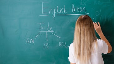 English teacher with long blonde hair at a chalkboard with grammar and words