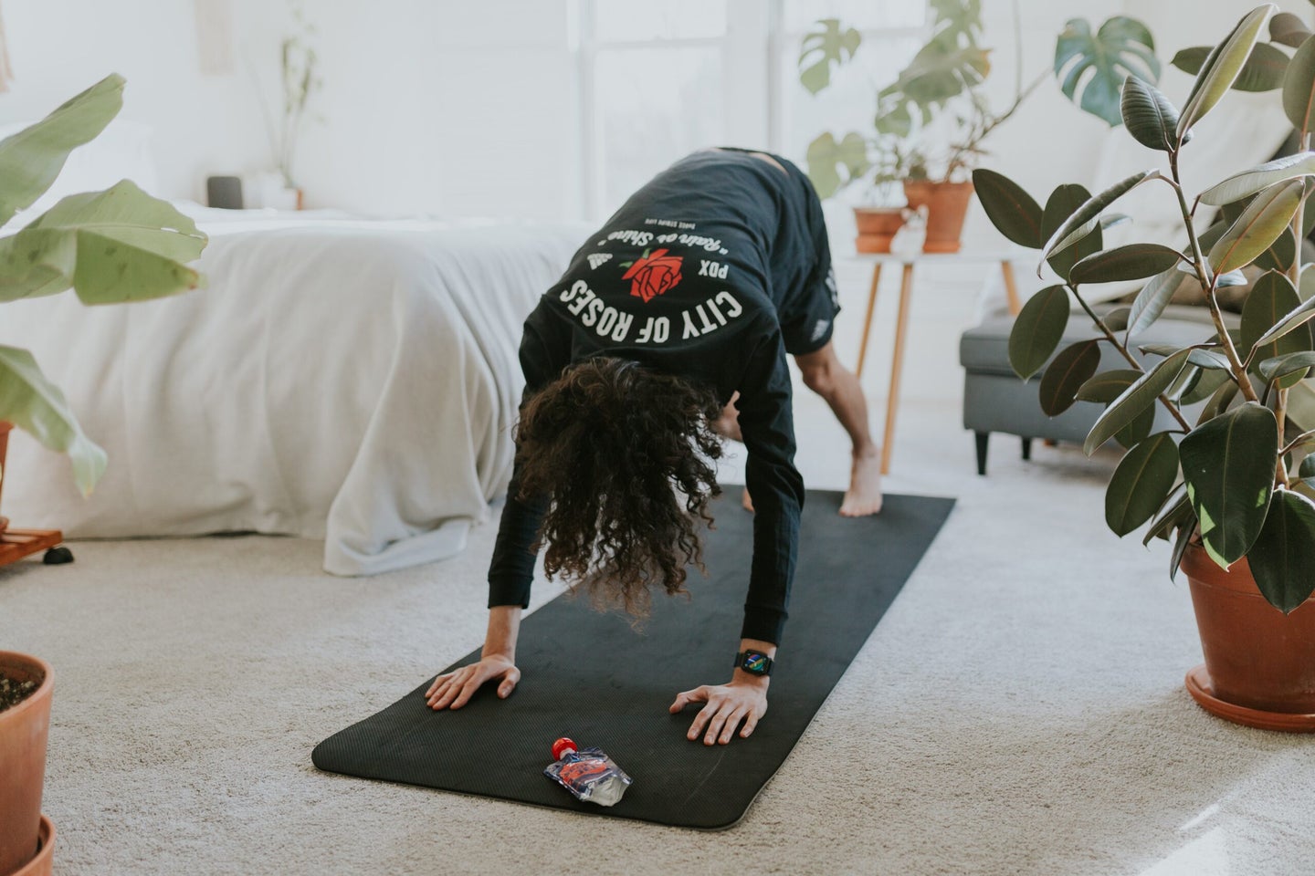 A person wearing a t-shirt and shorts doing yoga on a black yoga mat in a white bedroom near some plants.