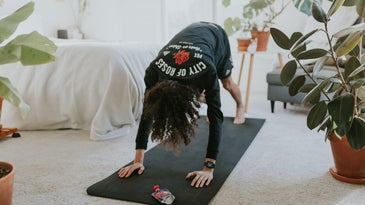 A person wearing a t-shirt and shorts doing yoga on a black yoga mat in a white bedroom near some plants.