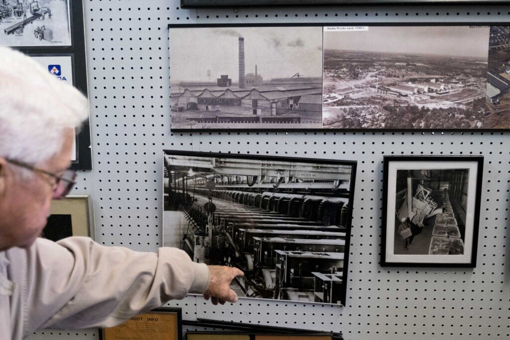 Badin museum curator with white hair, glasses, and tan shirt pointing to four black and white photos from the Alcoa aluminum plant