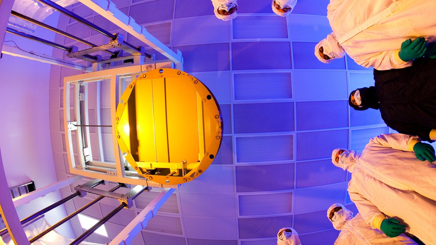 James Webb telescope primary mirror coated in gold seen from a fisheye view looking up with engineers in masks and protective gear looking down from a blue ceiling