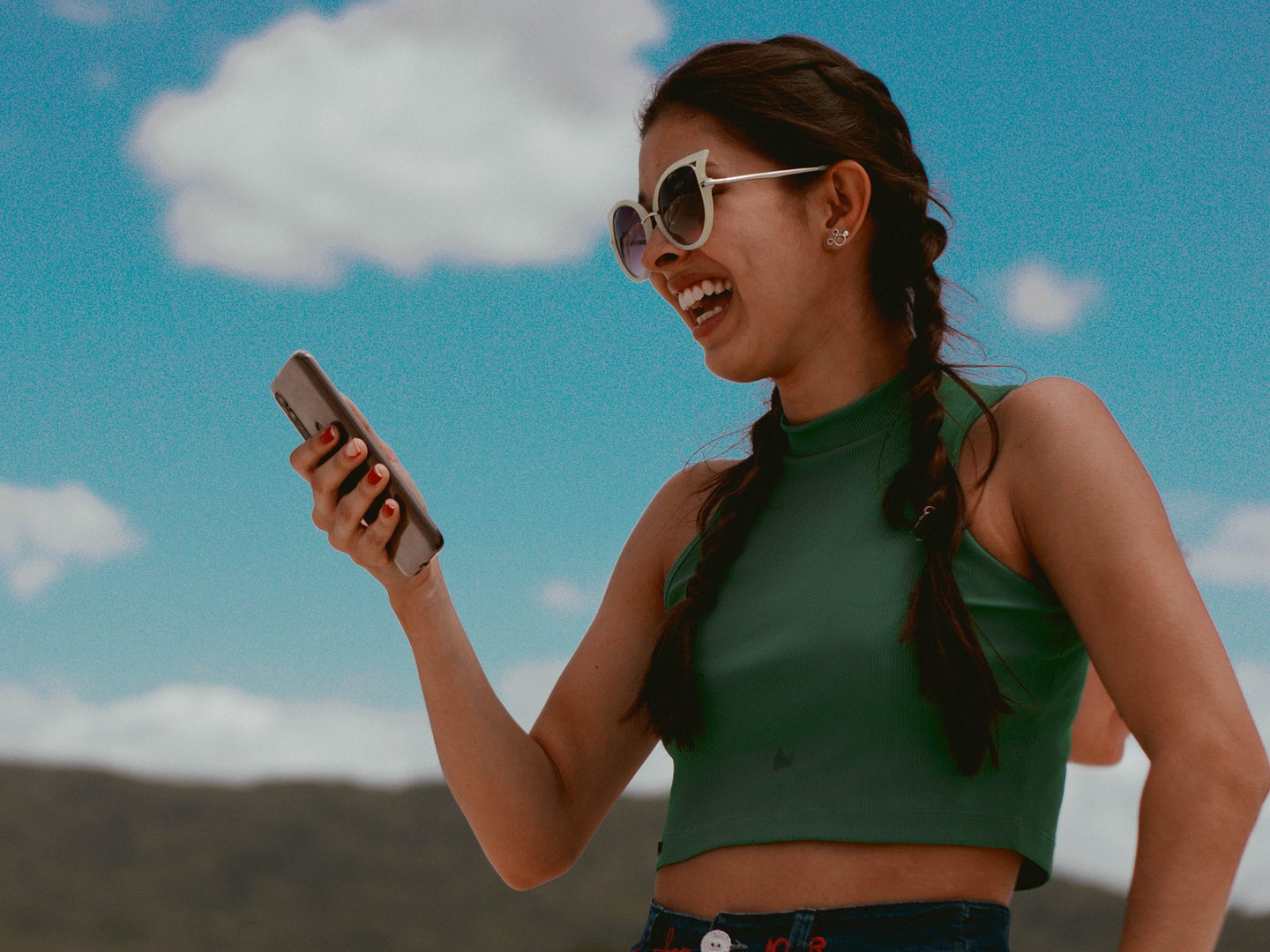 A woman with pigtails wearing sunglasses, holding a phone, and laughing under a blue sky.