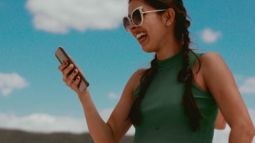 A woman with pigtails wearing sunglasses, holding a phone, and laughing under a blue sky.