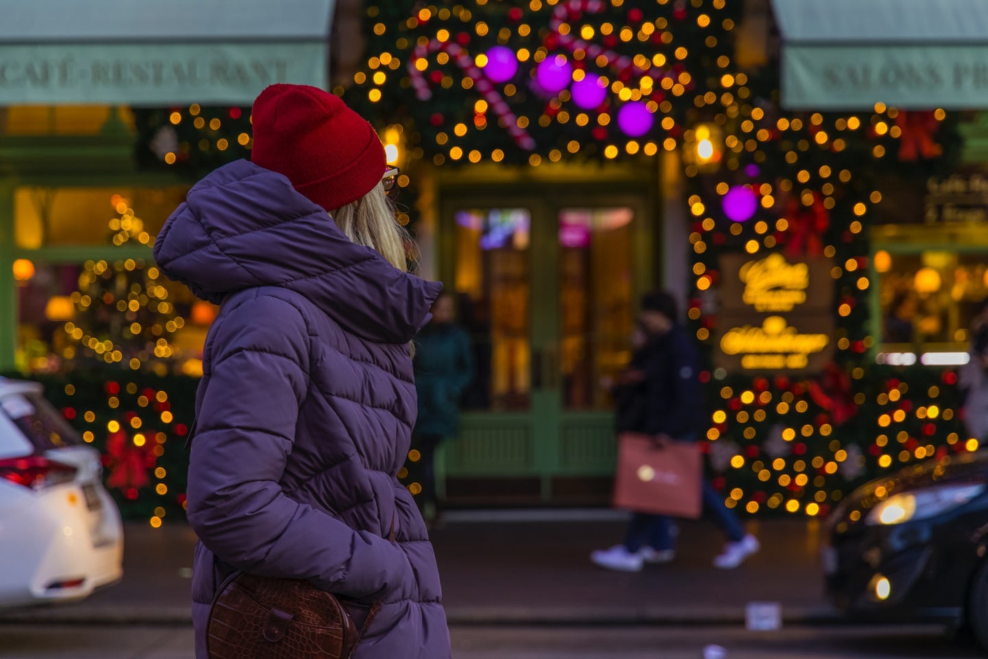 a woman in a purple coat and a red hat looks at a shop covered in christmas lights