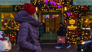 a woman in a purple coat and a red hat looks at a shop covered in christmas lights