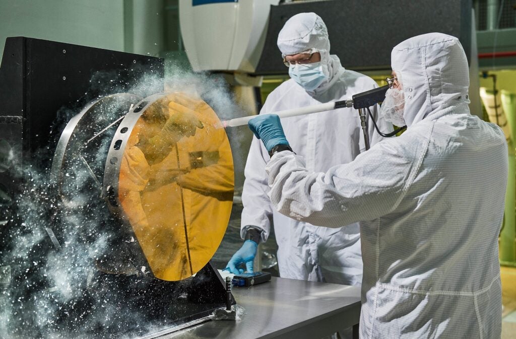 Engineers in protective gear blasting one of the James Webb Space Telescope's gold-coated primary mirrors with white powder
