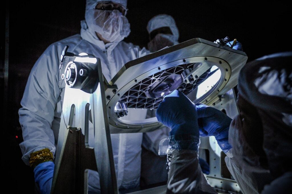 Two NASA engineers in protective clothing looking at micro sensors from the James Webb Space Telescope under a white light