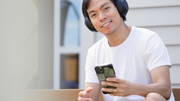 A man in a white t-shirt and black shorts sitting on a bench holding an iPhone and wearing black wireless headphones.