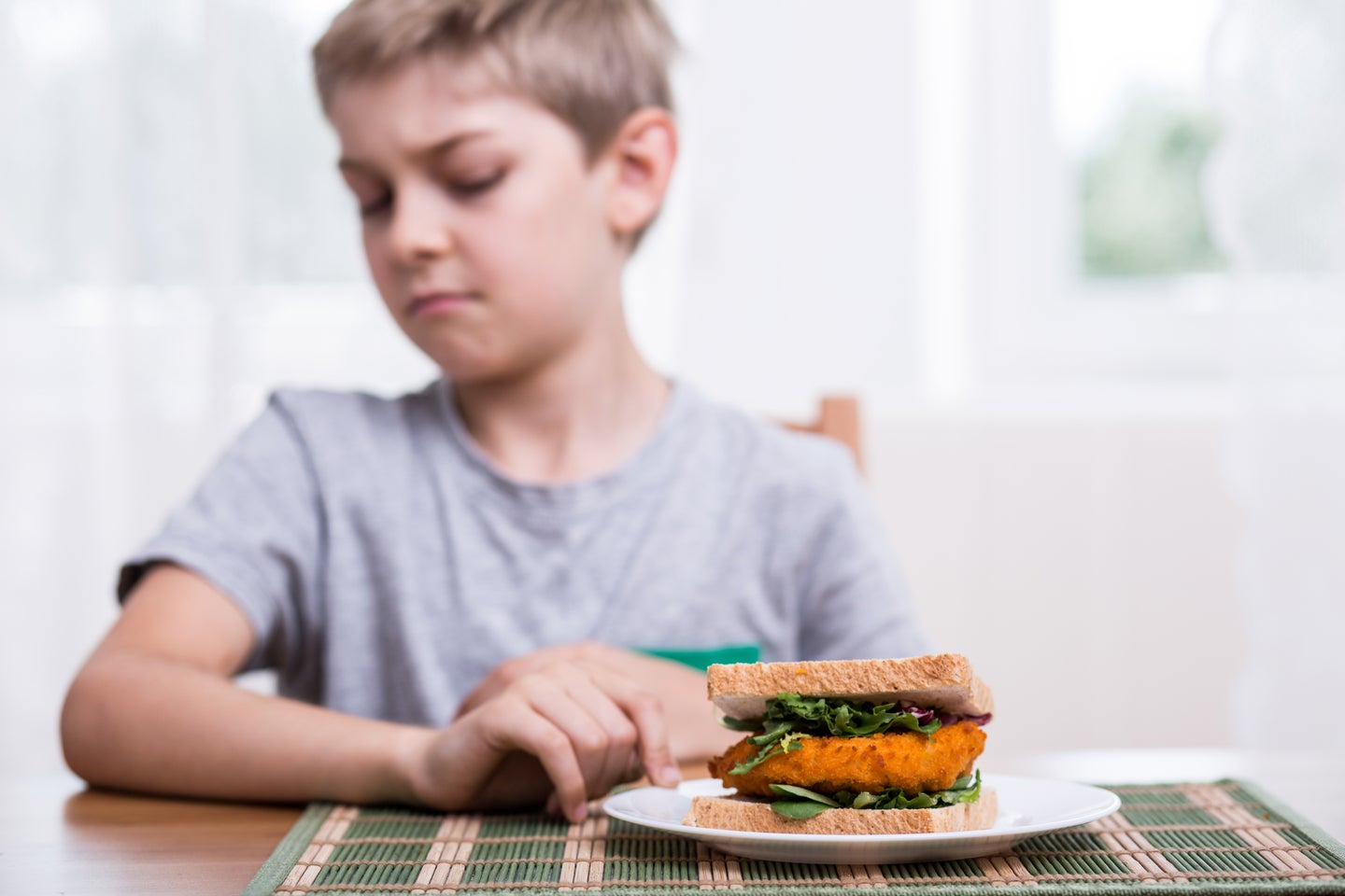 Kid with short blond hair and gray sports shirt turning away from a sandwich on a white plate on a wooden table because of a diet