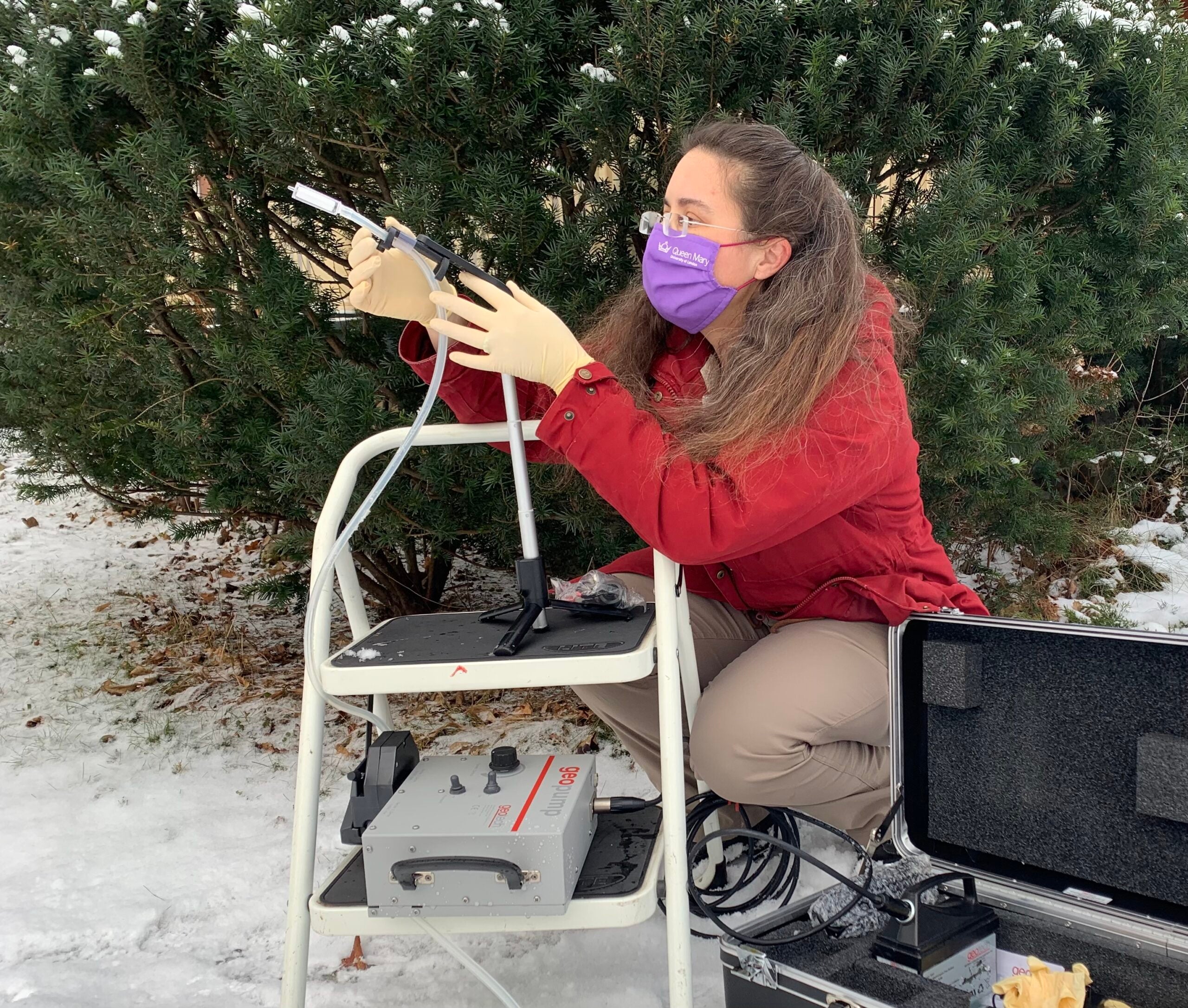 a woman in a purple face mask, gloves, and red jacket holds a piece of air collection equipment, which has a long tube feature. she is kneeling down next to a stool where the equipment is perched. behind her is a large green bush