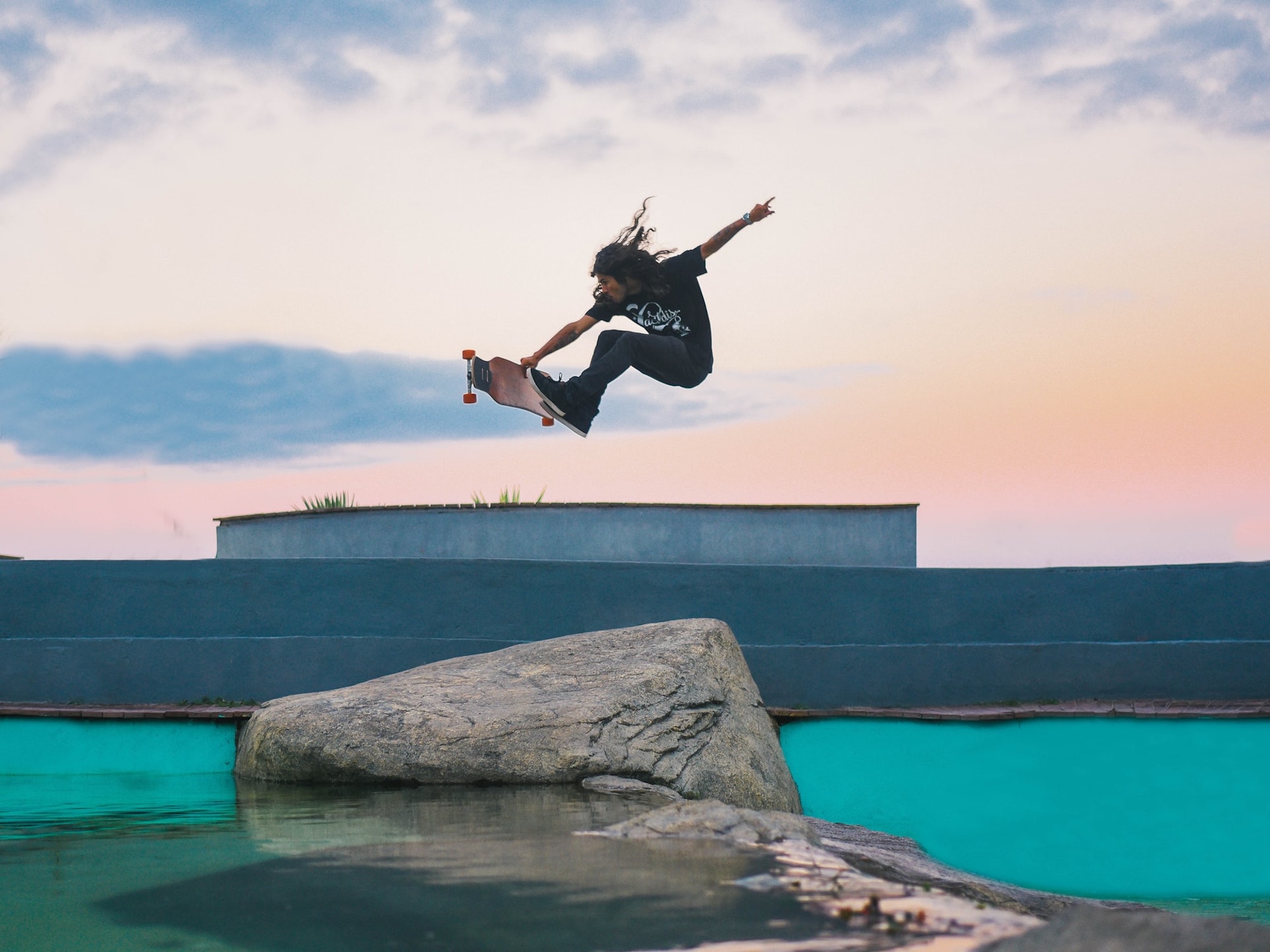 Skateboarder with long hair and black clothes doing a trick over a rock next to the sea green ocean