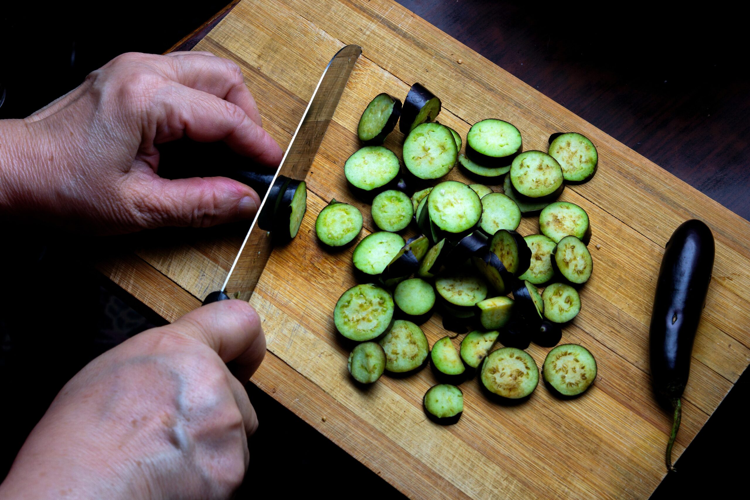 A person slicing cucumbers on a scratched wooden cutting board.