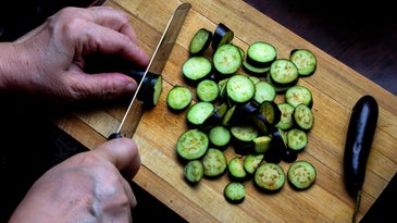 A person slicing cucumbers on a scratched wooden cutting board.