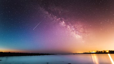 A shooting star in a purple, blue, and yellow night sky over water