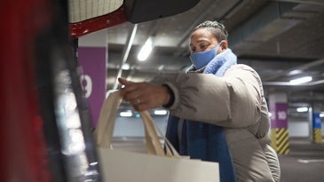 Brown-skinned person with long dark hair in a jacket and blue surgical mask getting into a car with a grocery bag during the Omicron COVID variant surge