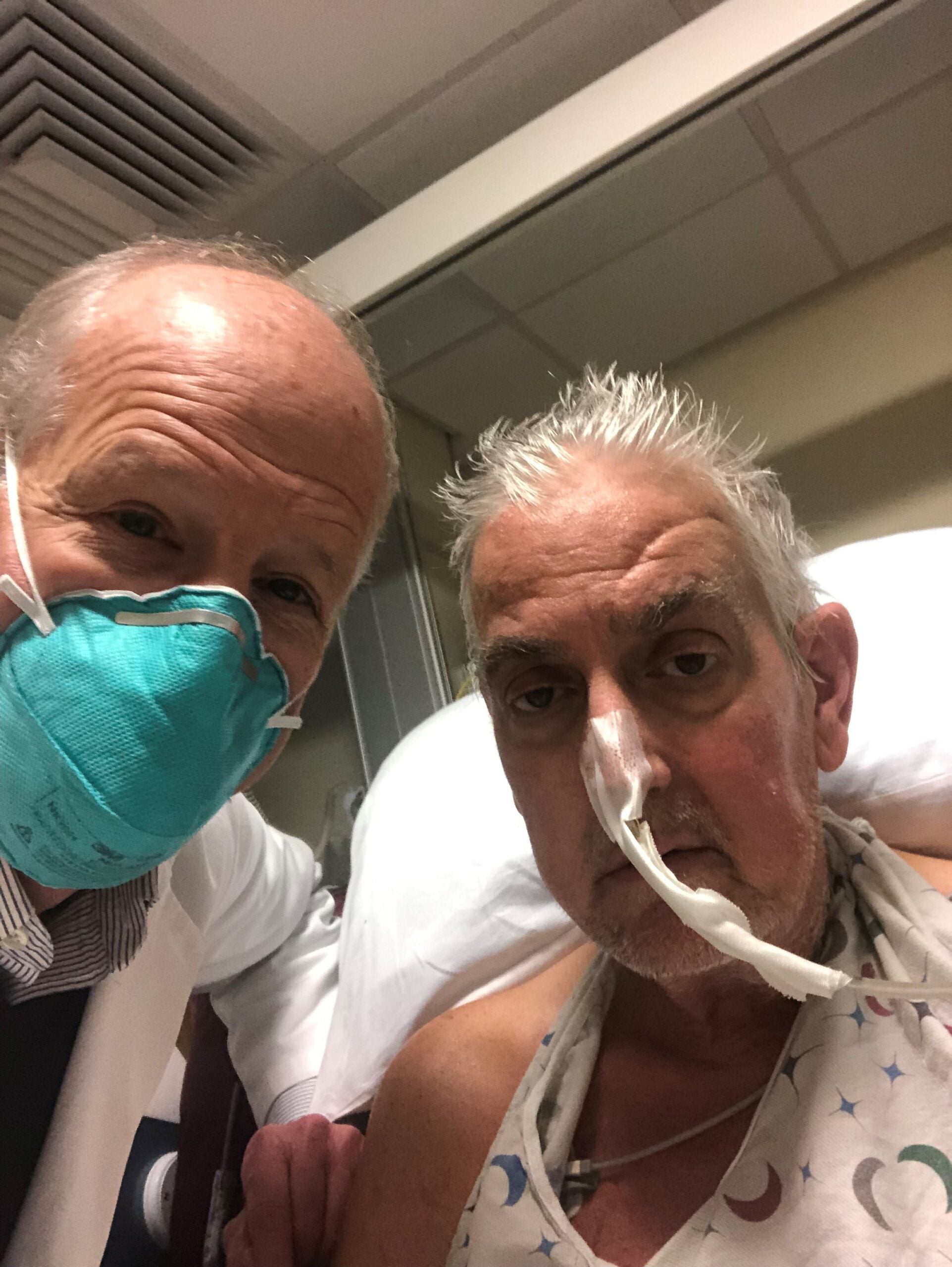 at a hospital room, a male surgeon with a blue medical mask on the left stands next to his patient, an older man sitting in bed in a gown with a oxygen tube hooked to his nose.