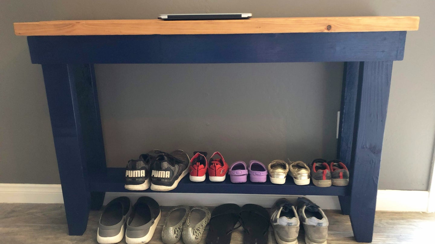 A blue wooden shoe rack table against a gray wall with neatly paired shoes on it.