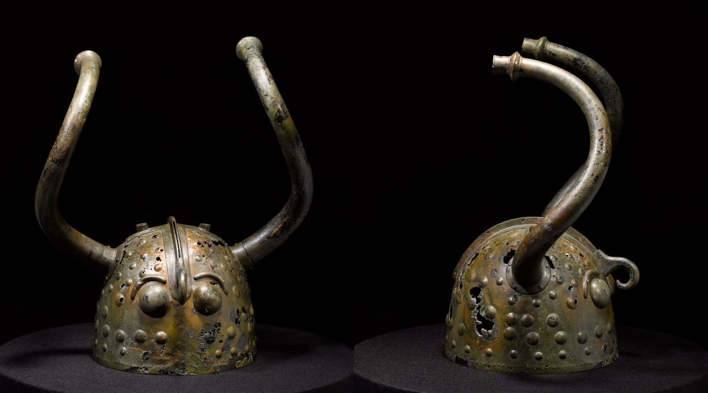 A bronze helmet shown from the front and the side. It has curving horns coming out of the sides of the head, and a birdlike face.