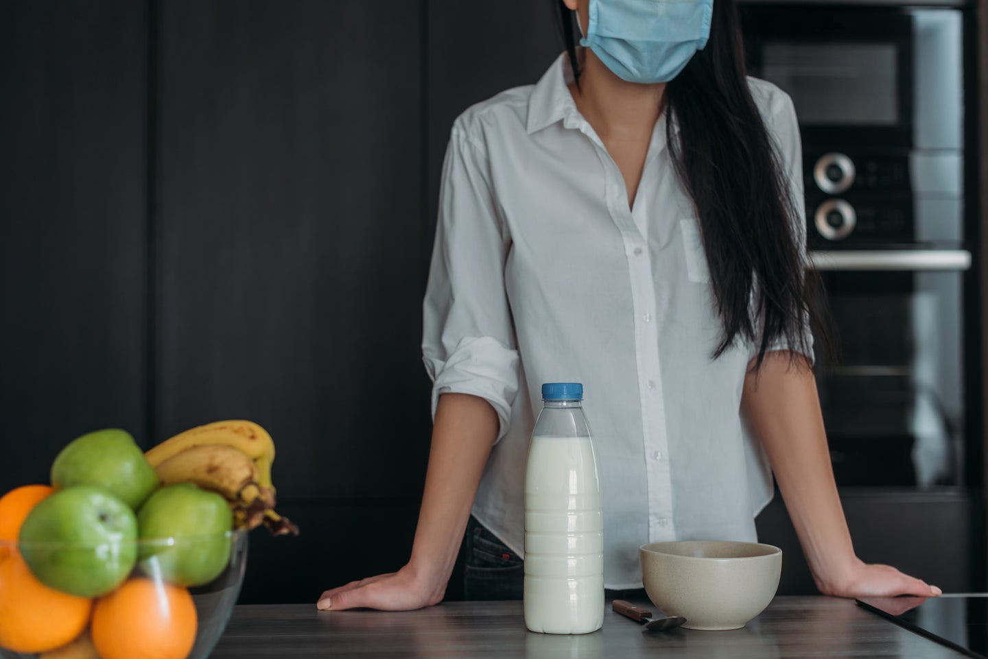 Person in white buttondown and with long black hair in COVID surgical mask leaning against a table with milk, a bowl of cereal, and green apples and oranges