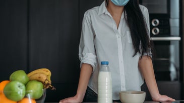 Person in white buttondown and with long black hair in COVID surgical mask leaning against a table with milk, a bowl of cereal, and green apples and oranges