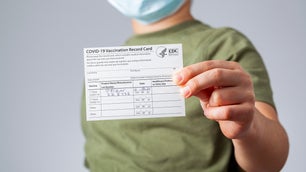 Kid in surgical mask and green shirt holding a COVID vaccination card because of a mandate