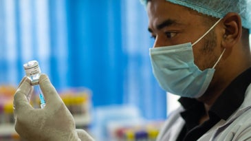 a doctor in a face mask, gloves, and hair net draws a vaccine with a syringe out of a vial