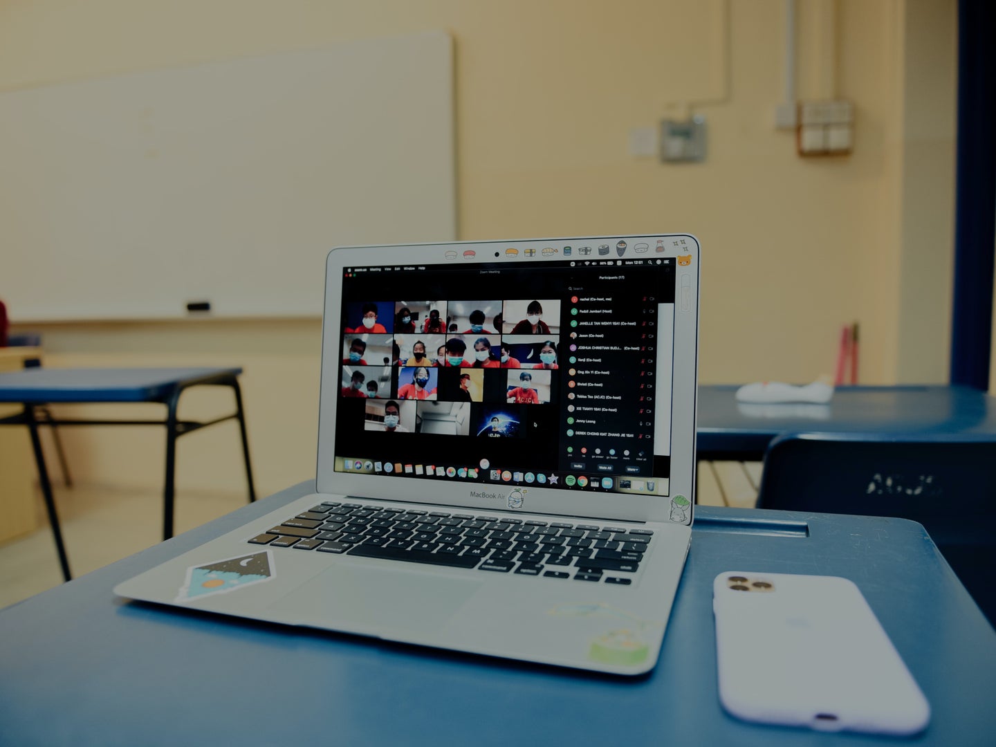 A laptop running Zoom sitting on a blue desk in an empty school classroom.
