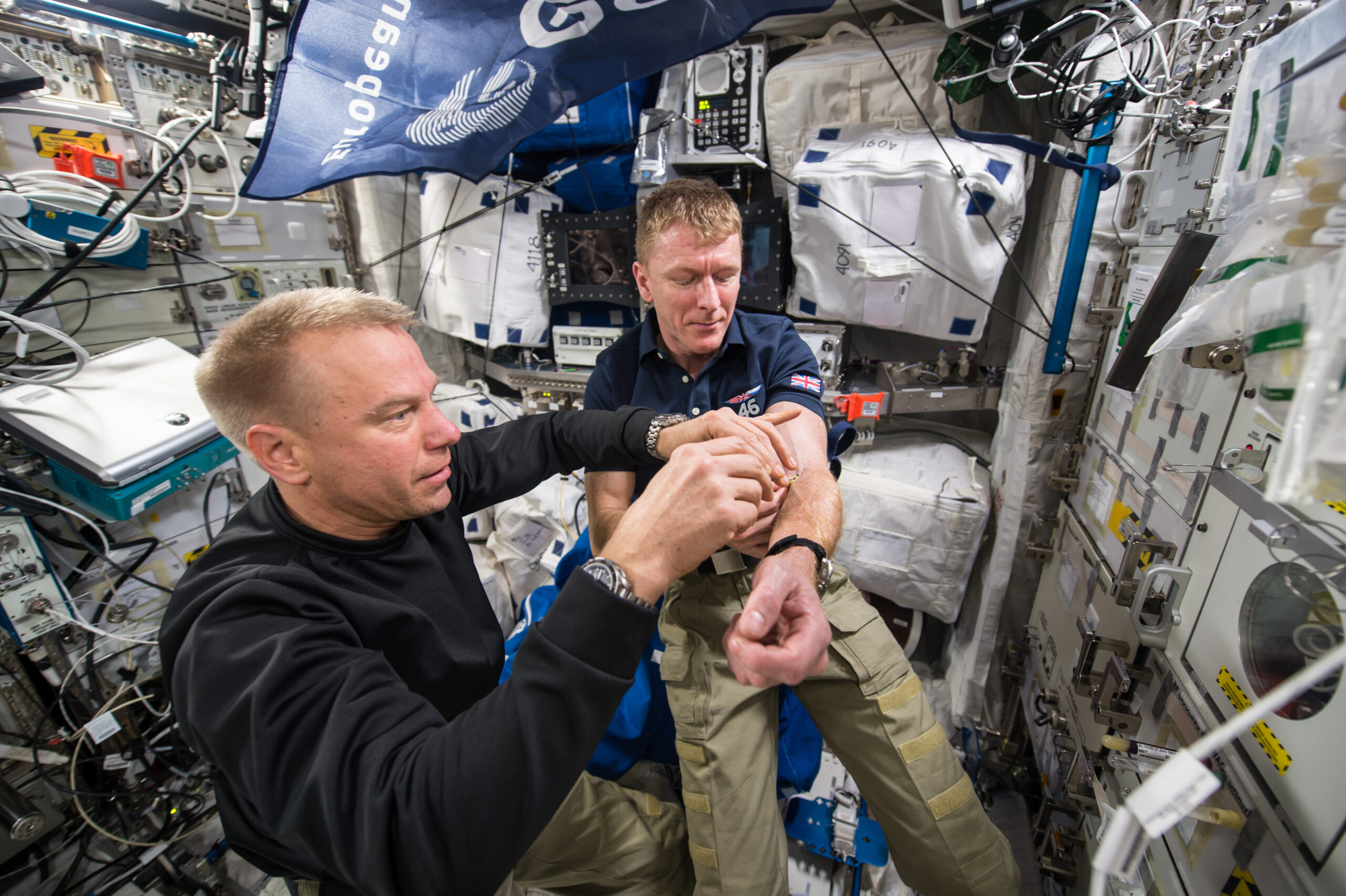 two male astronauts in a space station. one helps the other draw a blood sample from his arm