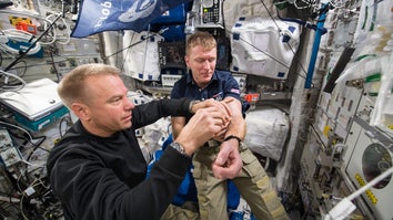 two male astronauts in a space station. one helps the other draw a blood sample from his arm