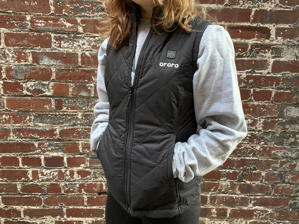 A woman wears a black heated vest and stands against a brick wall