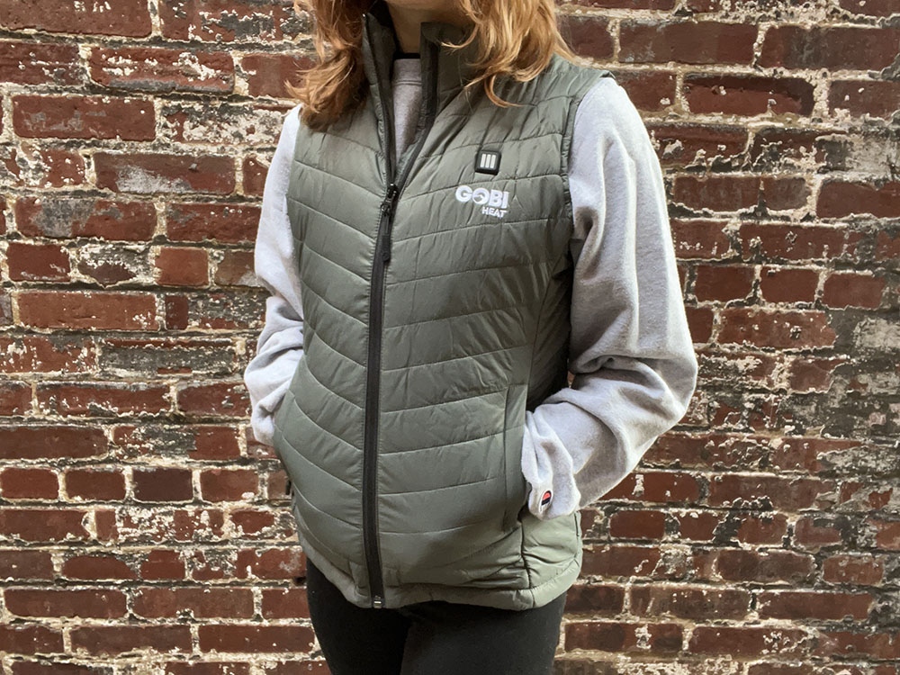 A woman stands against a brick wall wearing a sage green heated vest