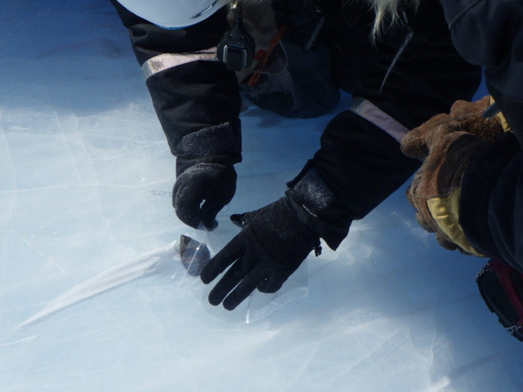 a scientist with winter gear and gloves places a meteorite in a plastic big while kneeling on a blue ice sheet