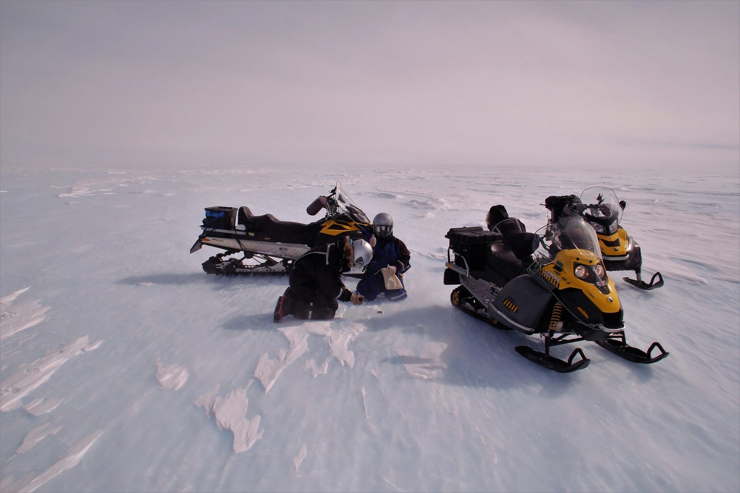 two researchers in winter gear crouch over a small rock in an icy windy landscape. next to them are yellow and black snow mobiles