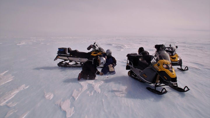 two researchers in winter gear crouch over a small rock in an icy windy landscape. next to them are yellow and black snow mobiles