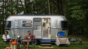 Black couple at a campfire in front of a silver Airstream RV trailer