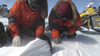scientists in big red winter coats, gloves and gear crouch down on an ice sheet to examine a dark gray rock embedded in the snow
