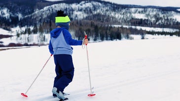 A child cross-country skiing on a snowy field near some round, tree-covered mountains.
