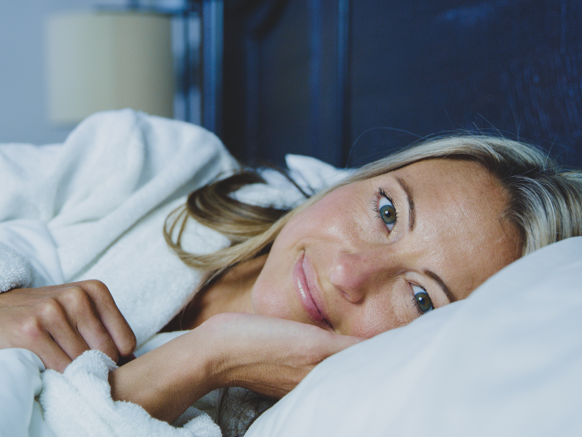 A blonde woman in a white bathrobe smiling and lying on a white bed in a blue room.