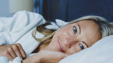 A blonde woman in a white bathrobe smiling and lying on a white bed in a blue room.