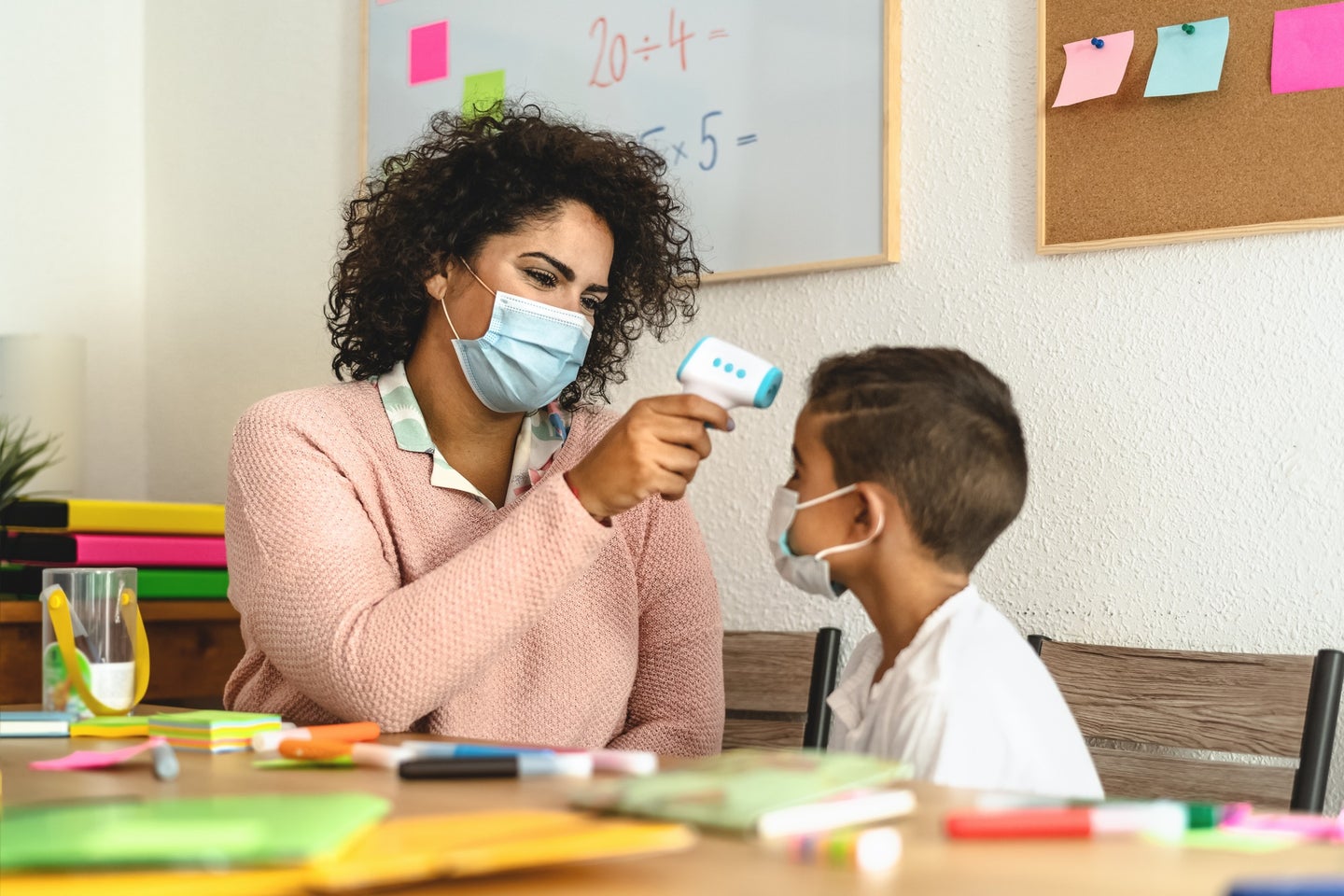 Daycare teacher in a surgical mask checking the temperature of a young kid in a surgical mask as a COVID precaution
