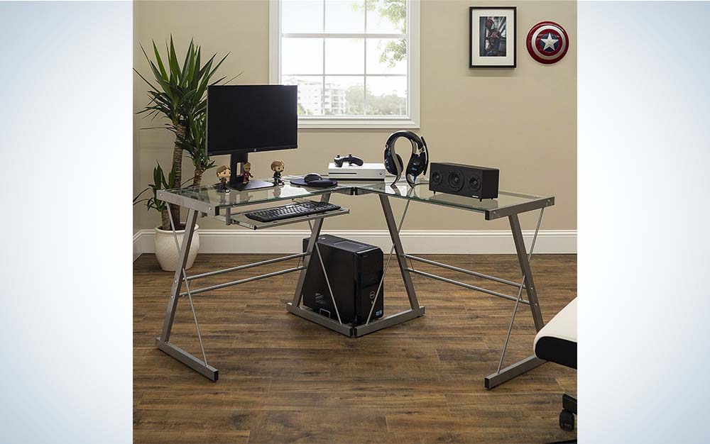 An L-shaped desk that's made of glass and metal in a room with a monitor on top and a CPU under it.