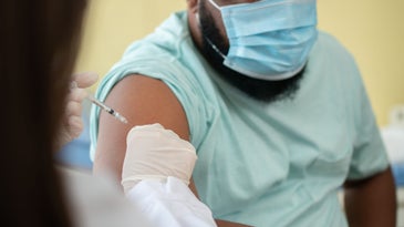 A man wearing a surgical mask is vaccinated by a healthcare worker wearing gloves.