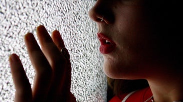 A woman with red lipstick on standing very close to a TV screen that's playing black and white static, as she gently touches the screen with her hand.
