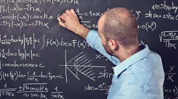 Man in blue shirt writing math onto a black chalk board.
