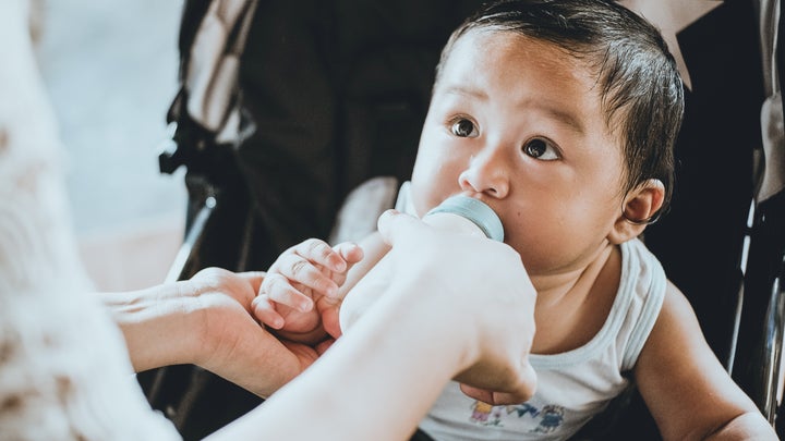 A baby in a stroller drinking formula from a bottle held by a woman.