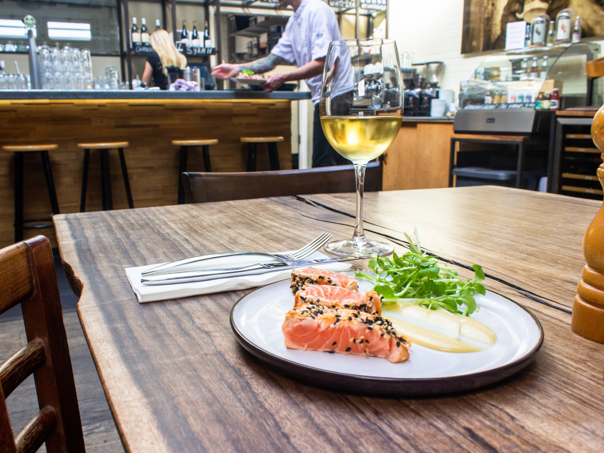 A cracked wooden table at a restaurant, with a glass of white wine and a white plate with salmon and greens on it.