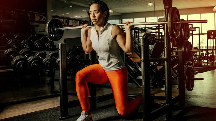 A woman in orange workout leggings and a gray sleeveless workout top doing a lunge squat with a barbell in a gym.