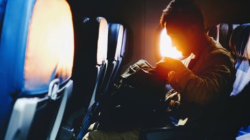 A man sitting on an airplane at sunset and looking into his backpack.